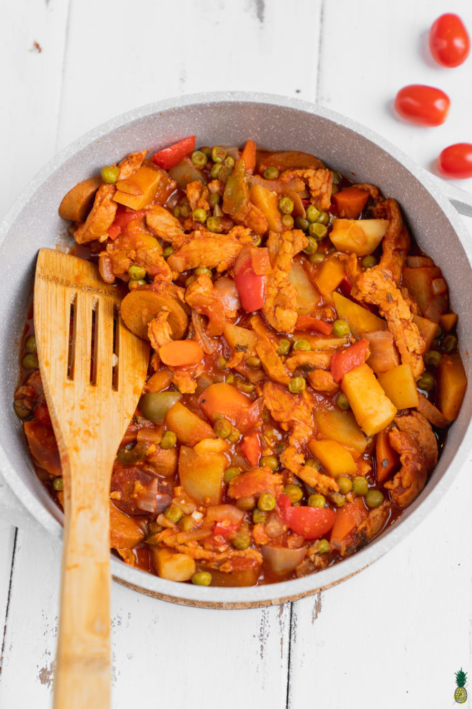 close up shot of a pan of vegan chicken afritada with a serving spoon and tomatoes