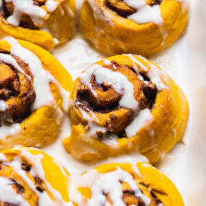 Close up shot of vegan pumpkin cinnamon rolls in a white dish with cream cheese icing drizzle by sweet simple vegan