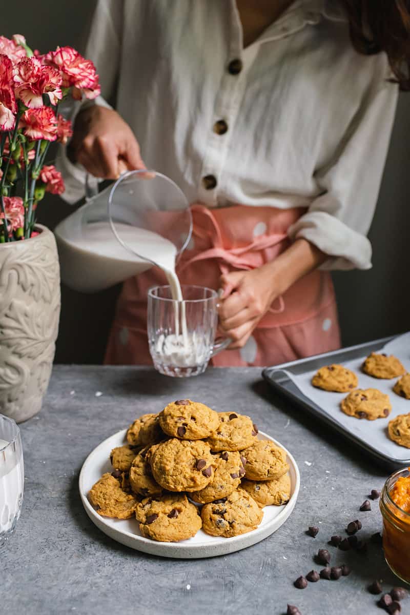 woman pouring milk into a glass behind a plate of pumpkin chocolate chip cookies