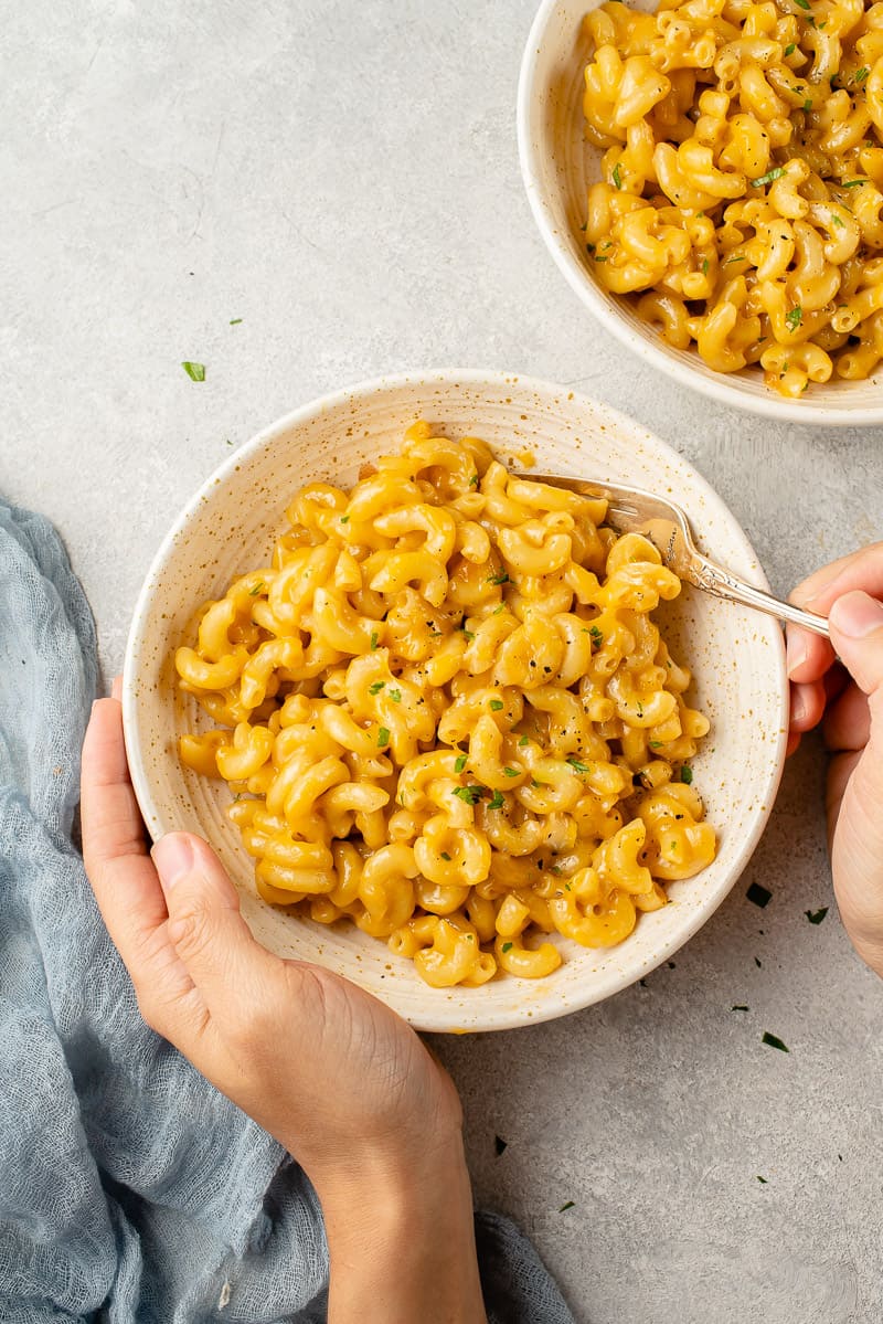 hands holding a white bowl of vegan mac and cheese with a fork