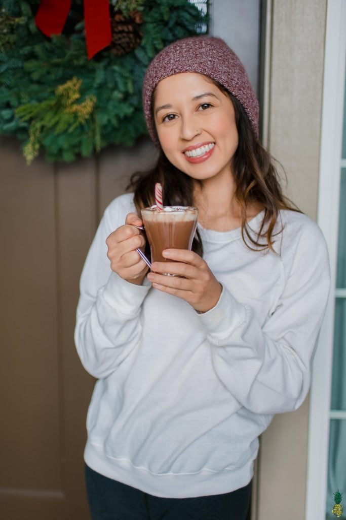girl in a red beanie posing with vegan hot cocoa with whipped cream in front of a christmas wreath; jasmine briones of sweet simple vegan