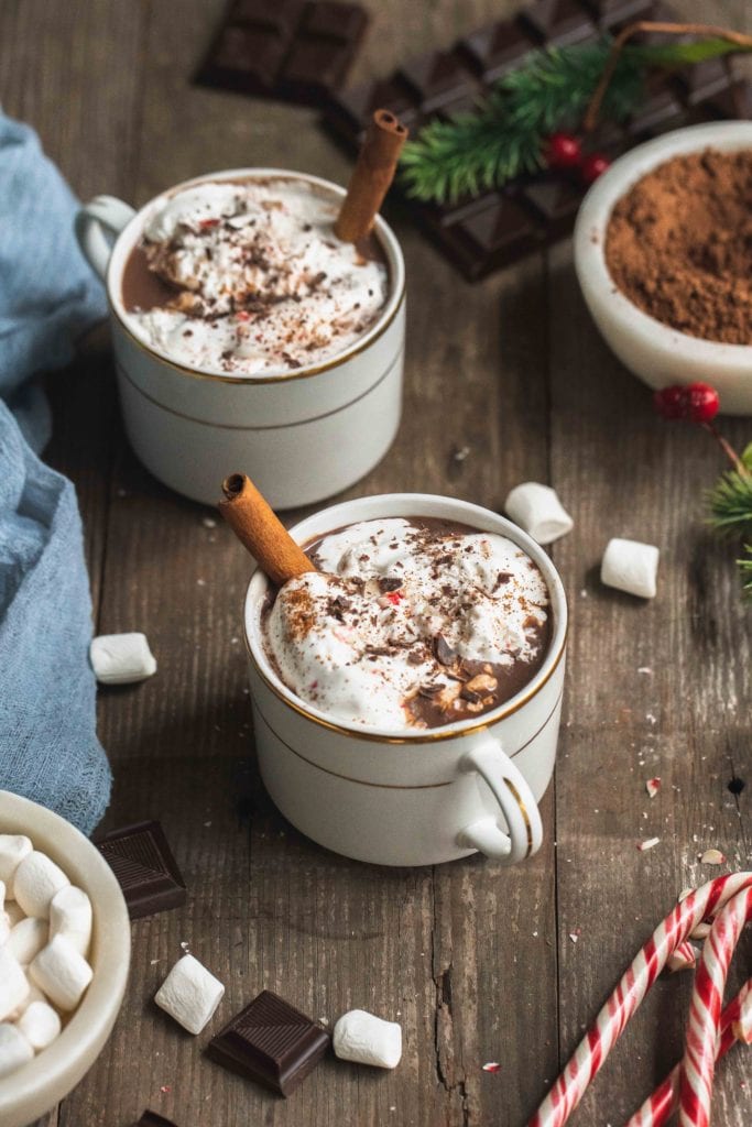 cups of vegan hot cocoa on a wooden board with whipped cream, peppermint and cinnamon sticks by sweet simple vegan