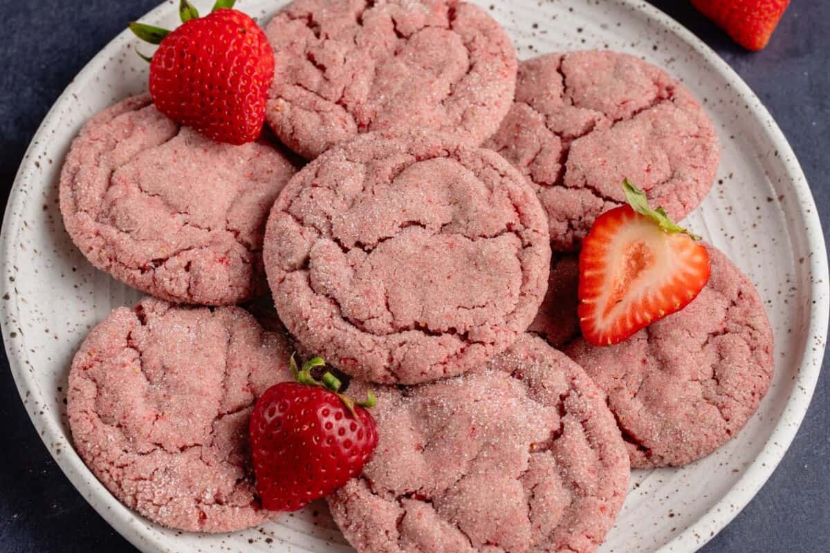 horizontal photo of strawberry sugar cookies stacked on a speckled white plate with fresh strawberry slices