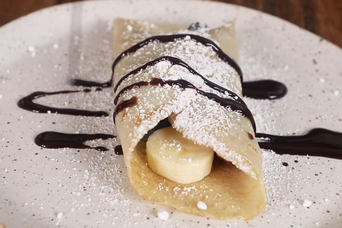 sweet crepe on white plate with fruit, powdered sugar and chocolate sauce