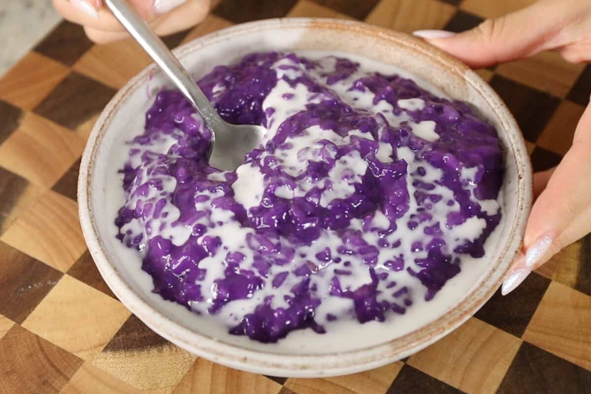 close up photo of spoon scooping ube champorado from white bowl on checkered board
