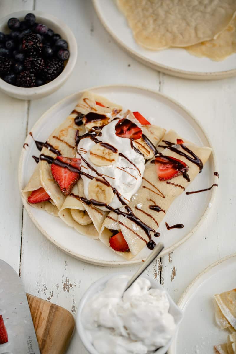 vegan crepes on a white plate with whipped cream, fresh fruit, chocolate syrup and powdered sugar