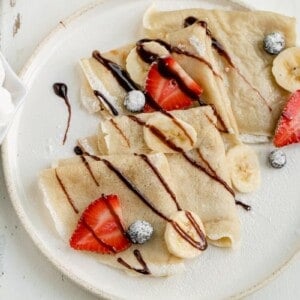 vegan crepes on a white plate with fresh fruit. chocolate syrup and powdered sugar